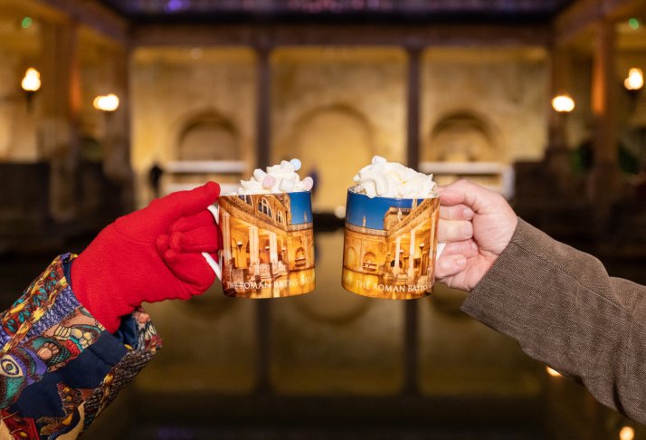 Image: Two people holding mugs of hot chocolate beside the Great Bath