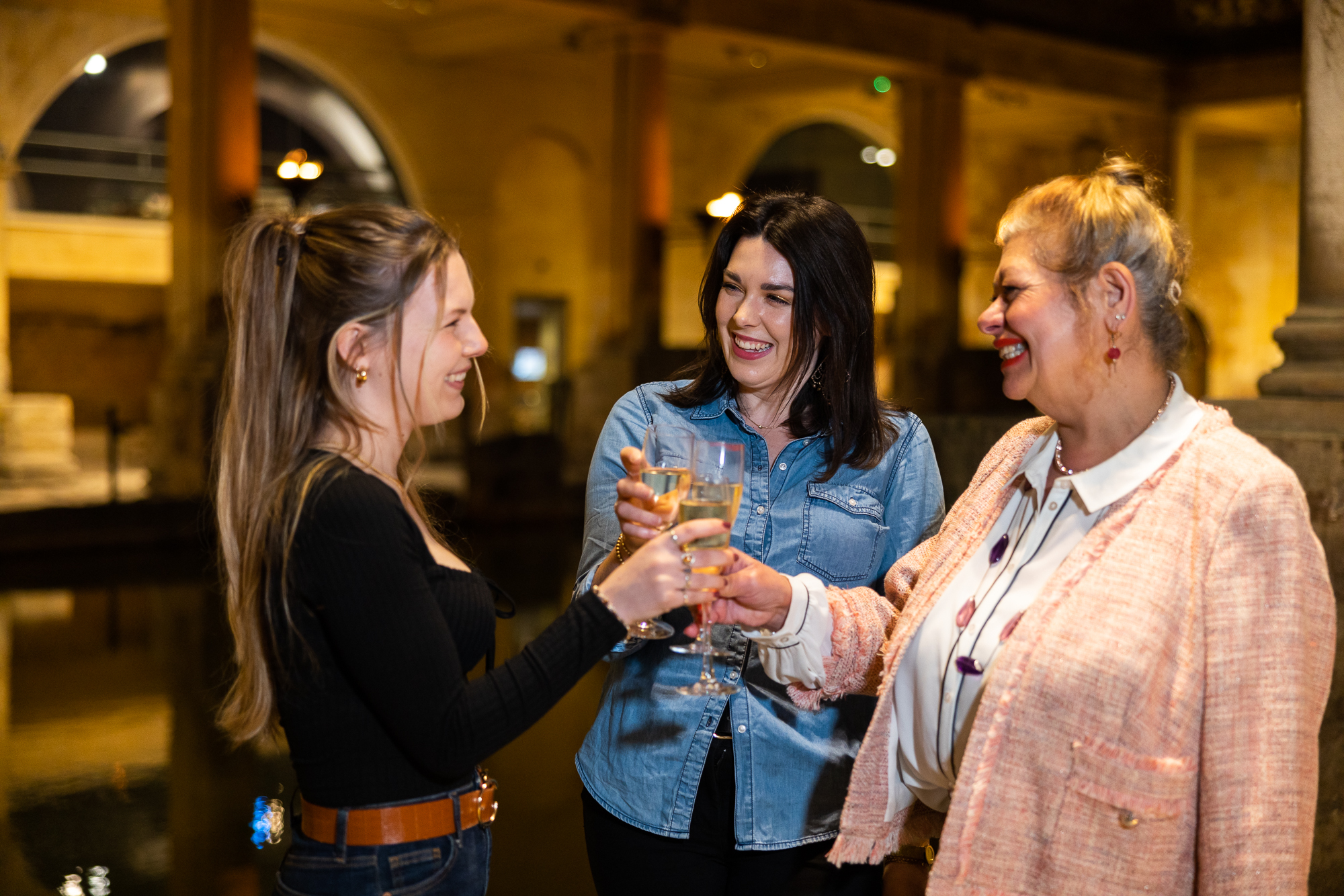 Image: Three visitors enjoying a drink beside the torchlit Great Bath