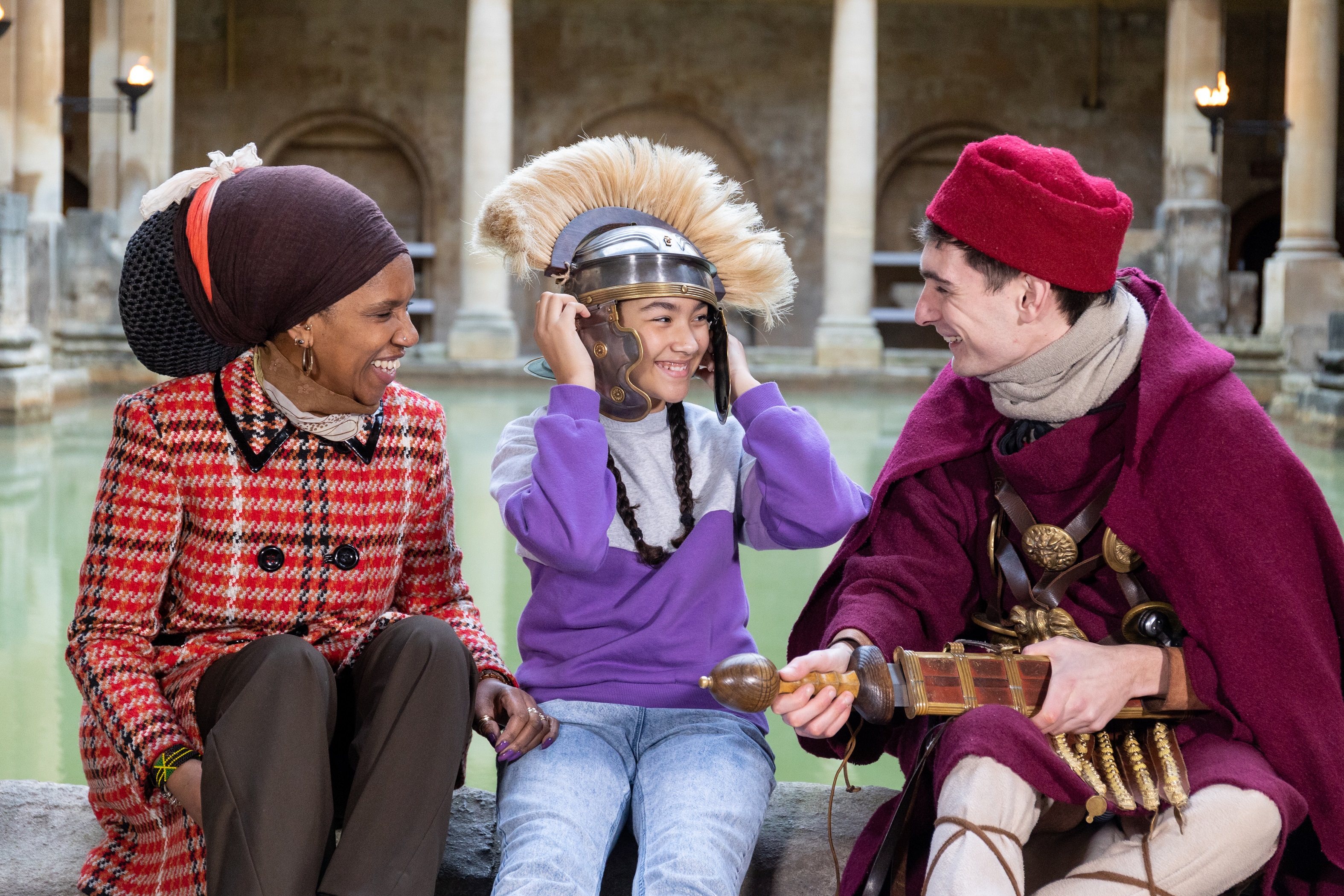 A girl trying on a Roman costumed character's helmet, beside the Great Bath