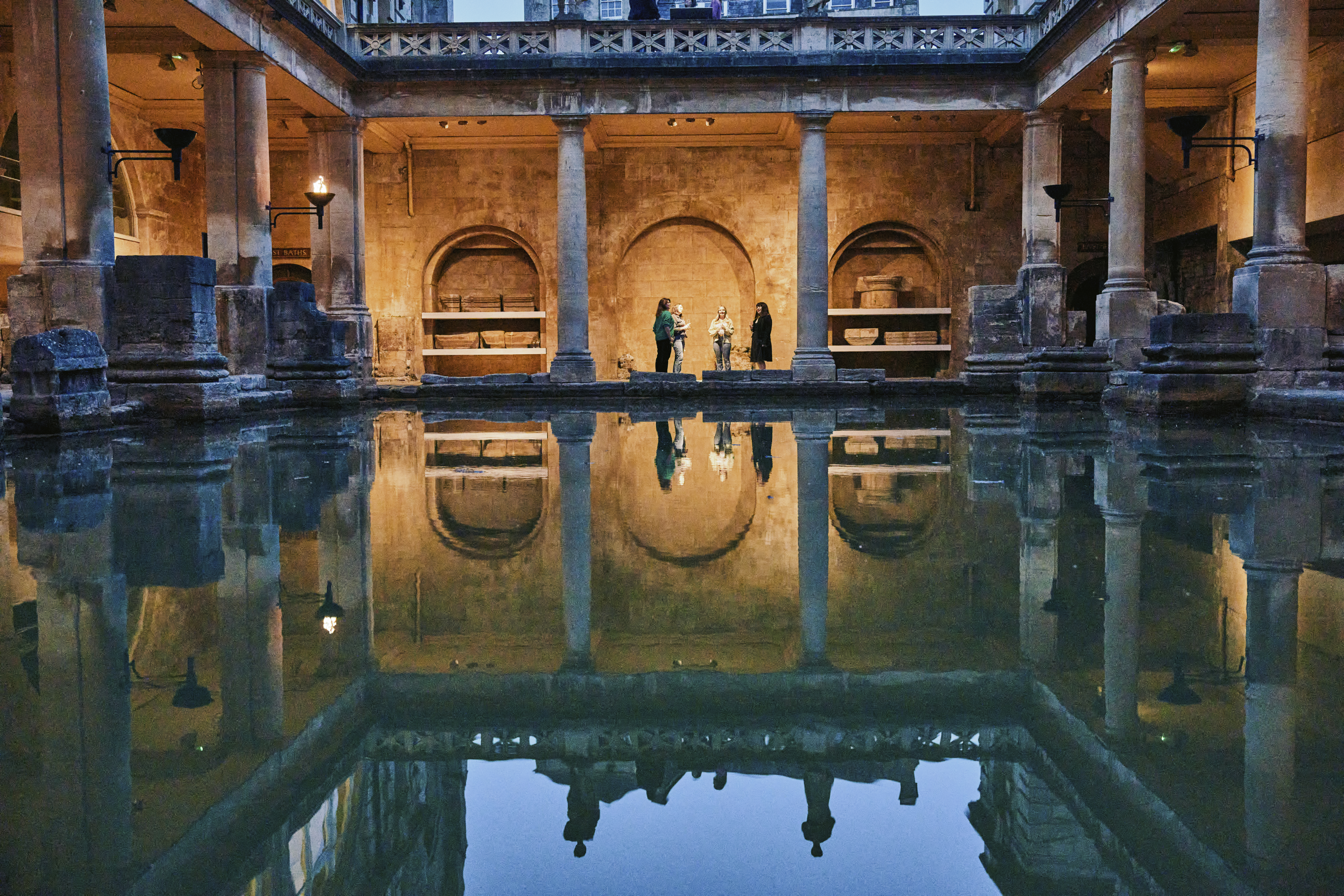 Roman Baths by evening under torchlight. Four people stand at the far end at the Bath, their reflection mirrors them in the water. Around the bath the torches are lit, casting a golden glow