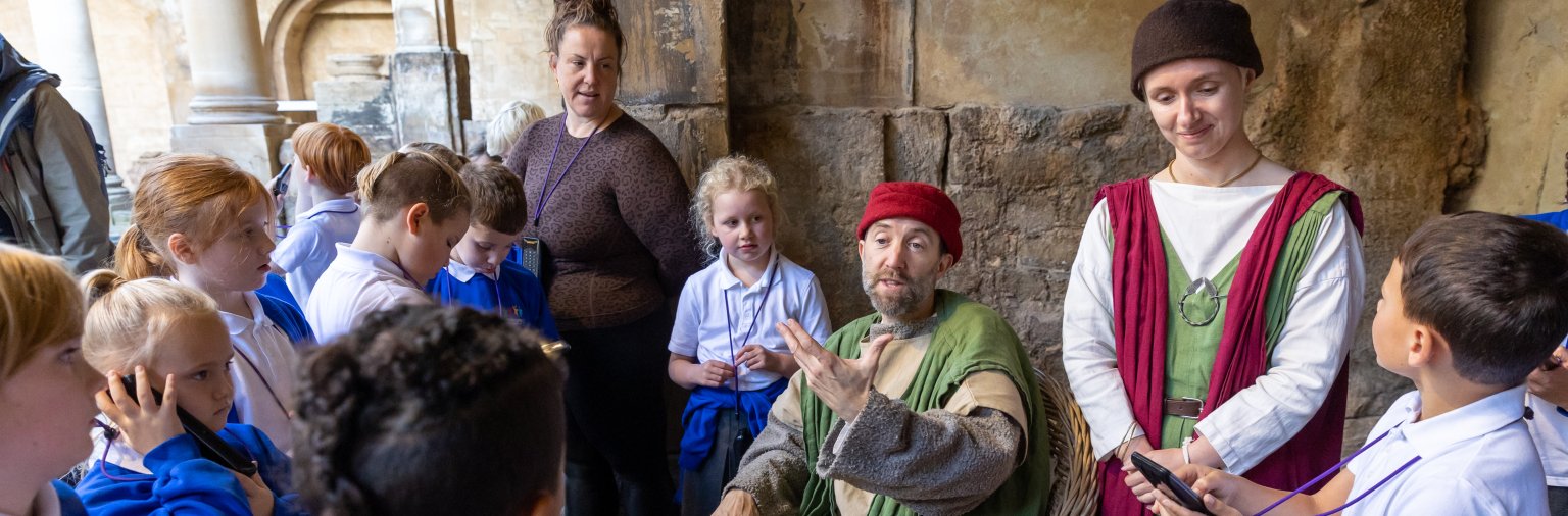 Image: A school group standing beside the Great Bath with a costumed character