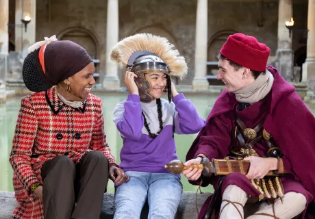 A girl trying on a Roman costumed character's helmet, beside the Great Bath