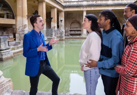 A male member of staff giving a tour beside the Great Bath, with four visitors listening.