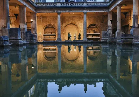 Roman Baths by evening under torchlight. Four people stand at the far end at the Bath, their reflection mirrors them in the water. Around the bath the torches are lit, casting a golden glow